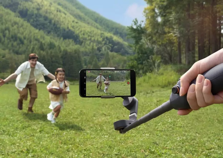 A person holds a DJI Osmo Mobile 6 gimbal with an extended selfie stick, capturing a video of a young girl running with a football while an adult playfully chases her in a lush green field. The smartphone mounted on the gimbal displays the live recording, showing the moment in action. The background features rolling hills, trees, and a bright outdoor setting.